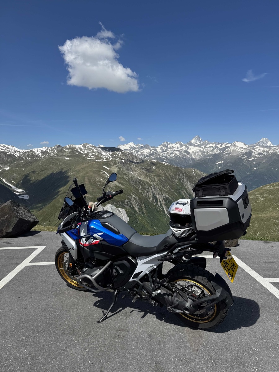 BMW adventure motorbike with panniers and top box parked at a high Alpine pass viewpoint, snowcapped mountains under a blue sky