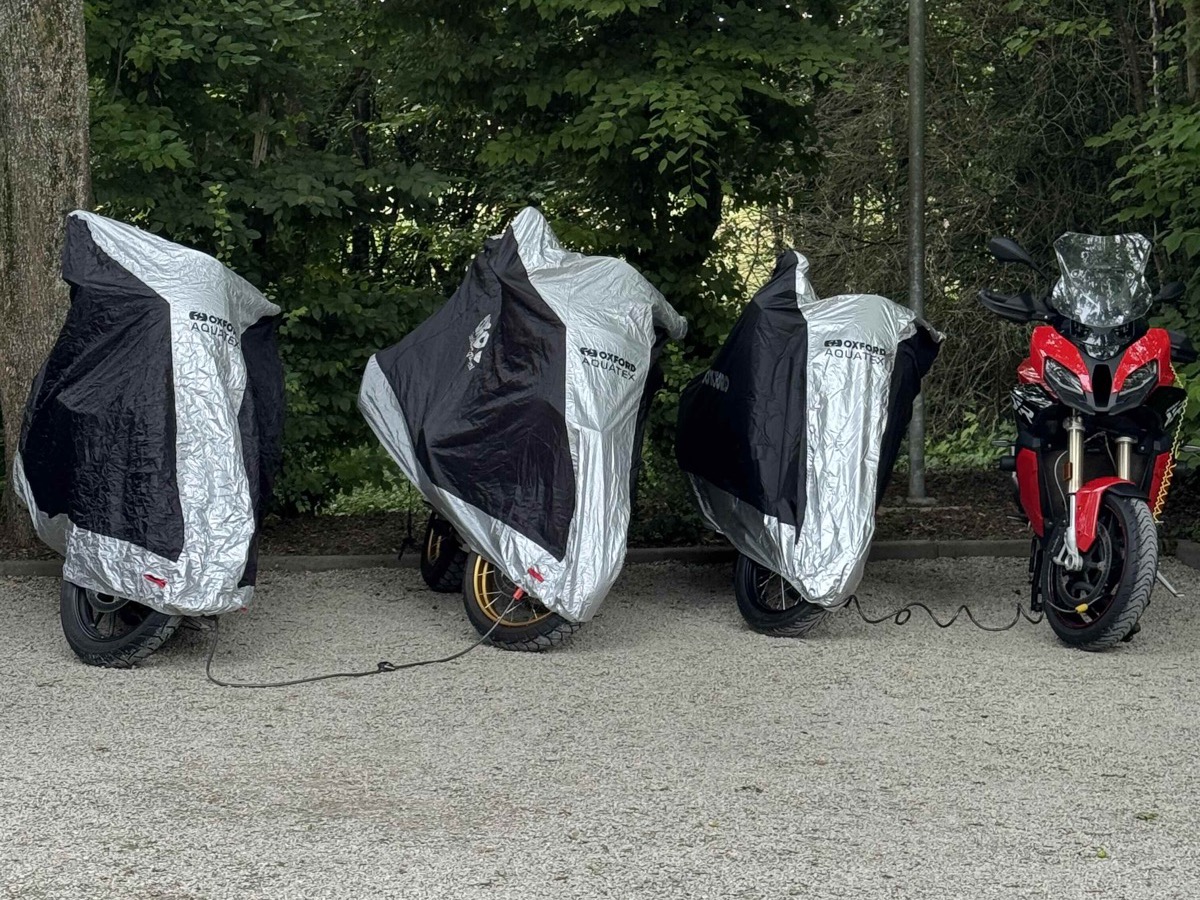 Four motorbikes parked for the night at Le Clos de Mutigny — three under silver-and-black covers and a red Ducati Multistrada uncovered, tall trees and an ornate lamp post in the background