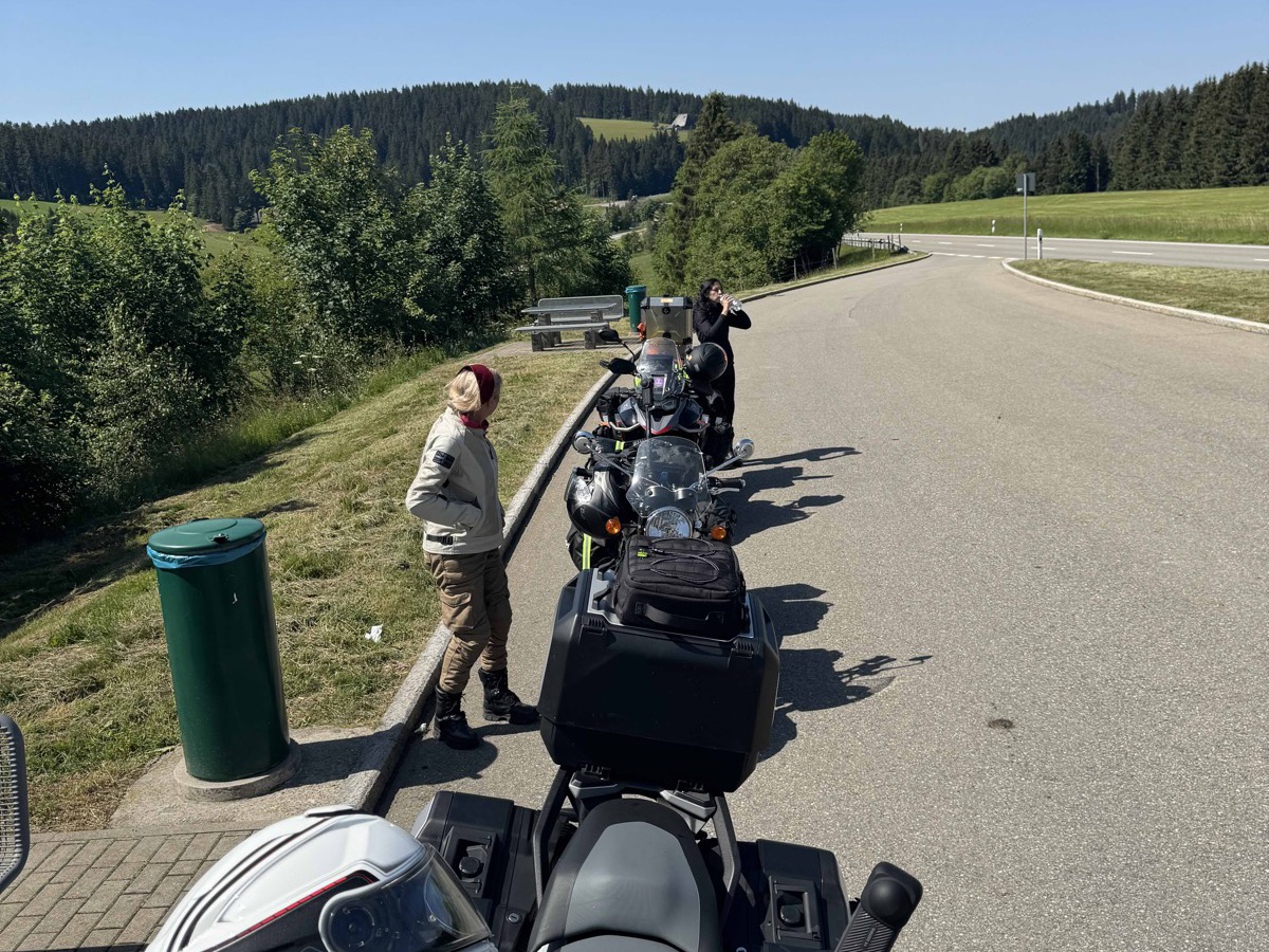 Four motorbikes lined up single-file at a quiet roadside pitstop on the long ride west — riders milling around, a small rest area with a green bin, country road and rolling green forested hills under a clear blue sky