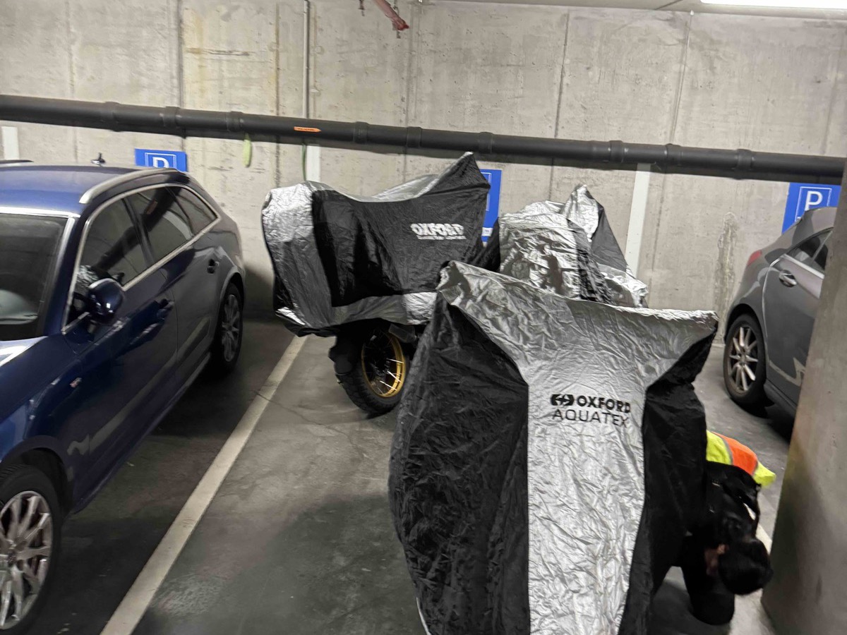 Four motorbikes parked together in a single bay (number 108) of an underground car park in Ghent, all covered with silver Oxford Aquatex motorbike covers, ordinary cars parked in the bays either side