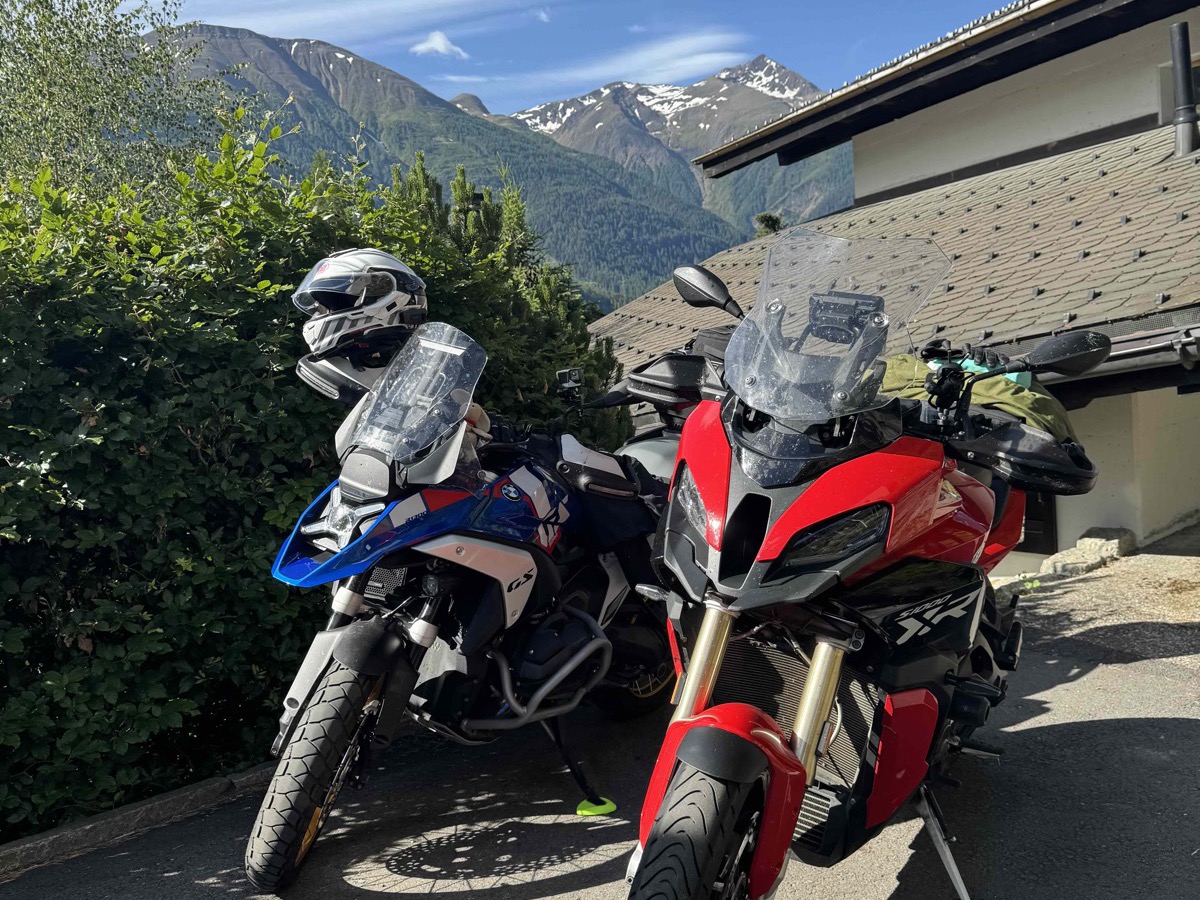 Two motorbikes parked at Chalet Arnica in Fiesch — a blue BMW GS adventure and a red Ducati Multistrada, snowcapped Alpine peaks rising behind them under a bright blue sky