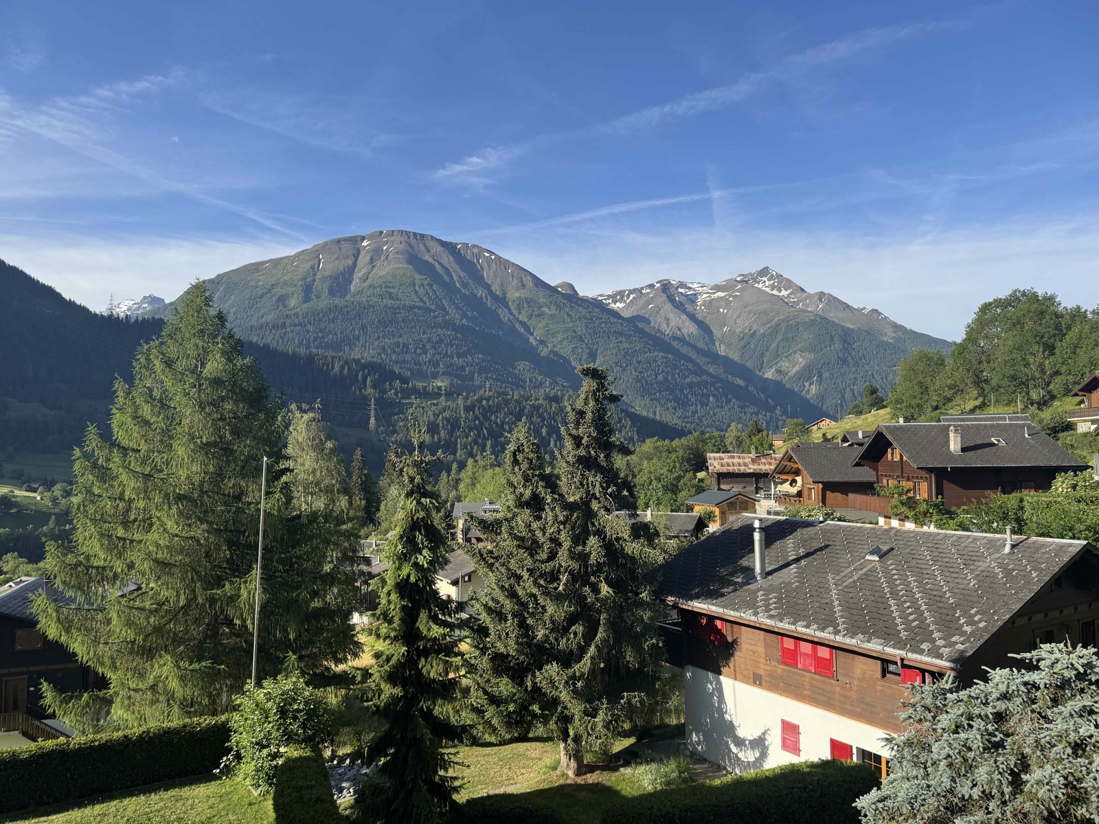 View from the Chalet Arnica balcony across Fiesch — neighbouring chalets in the foreground, a forested green peak rising in the middle distance, and snowcapped Alpine summits to the right under a clear blue sky