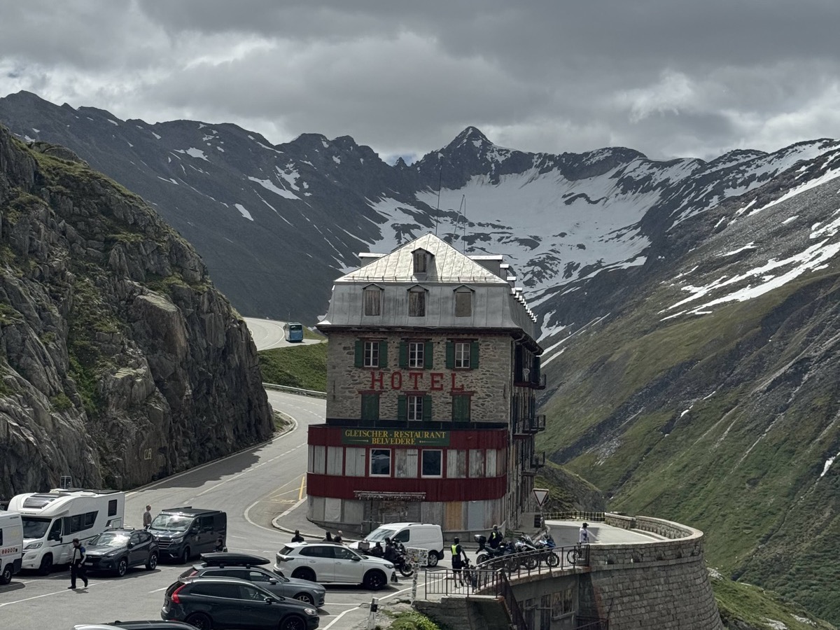 The historic Hotel Belvédère on the Furka Pass — a tall yellow-and-cream hotel building perched on a hairpin bend, snow-capped Alpine peaks rising behind under a moody grey sky, motorbikes and cars parked in the foreground