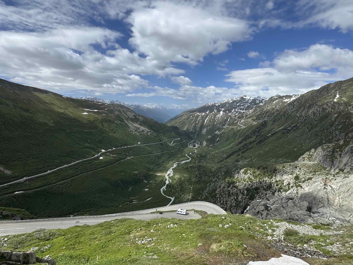 Looking down the Furka Pass road as it snakes through a green Alpine valley between two rocky ridges, patches of snow on the upper slopes and clouds drifting across a bright blue sky