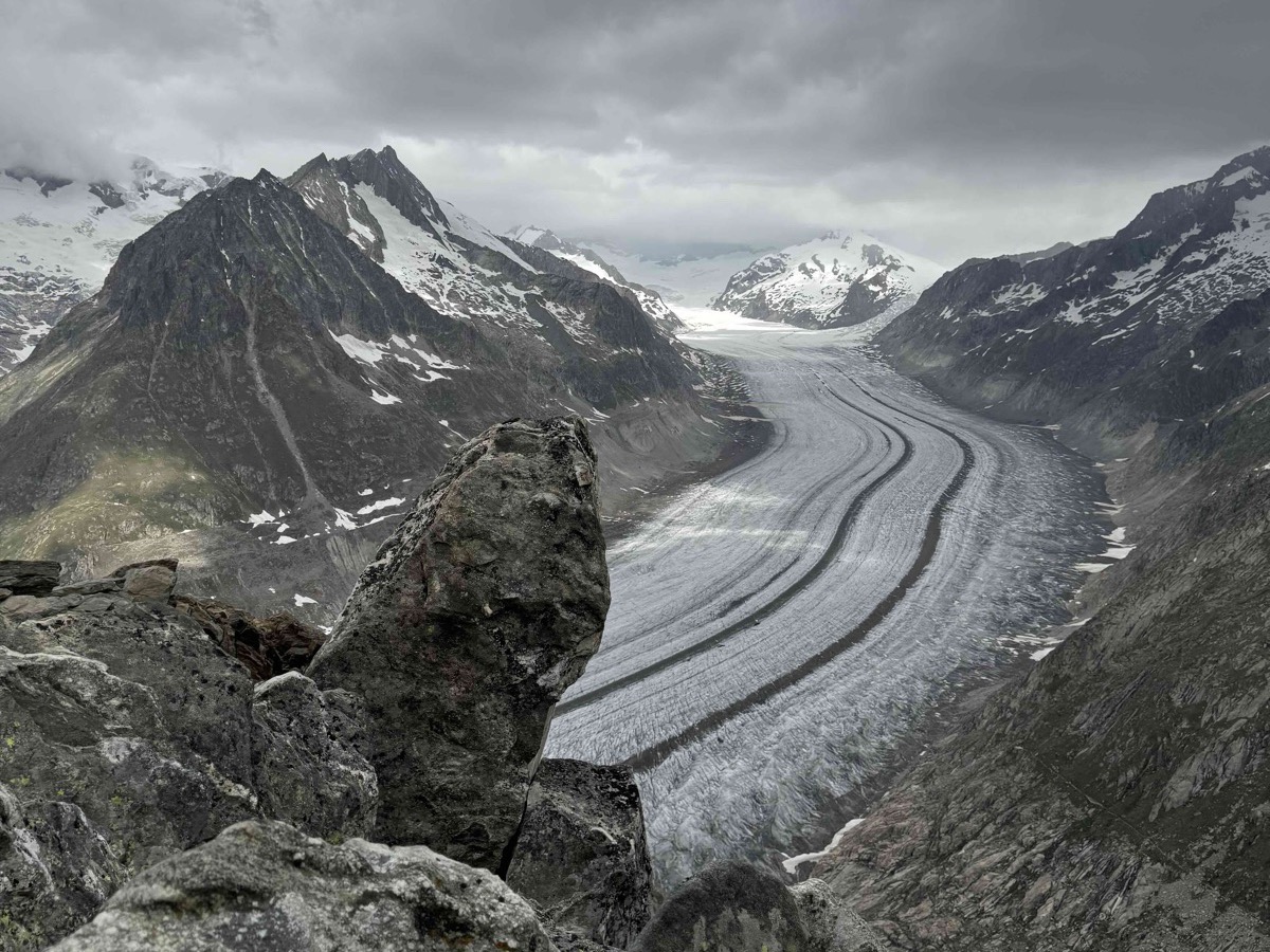The Aletsch Glacier seen from a rocky viewpoint above Fiesch — a vast curving river of grey-white ice running through a deep valley between dark Alpine ridges, snow-capped peaks at the head of the glacier under a moody grey sky