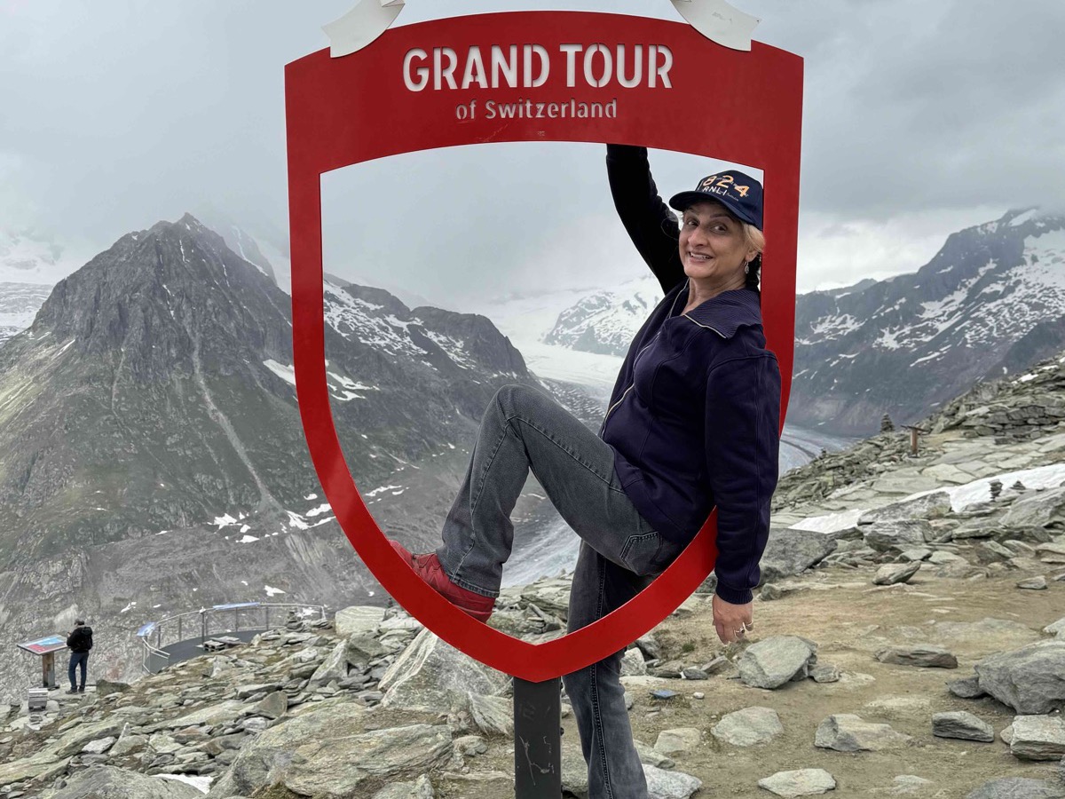 A rider posing in the red shield-shaped 'Grand Tour of Switzerland' photo frame at the Aletsch viewpoint, with the glacier and dark Alpine peaks framed behind under an overcast sky