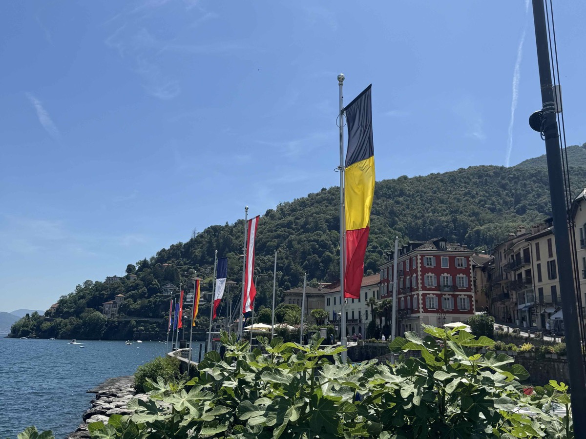 Lakeside promenade on Lago Maggiore — a row of flags including the Belgian tricolour, the lake and forested mountainsides to the left, Italian buildings to the right under a bright blue sky