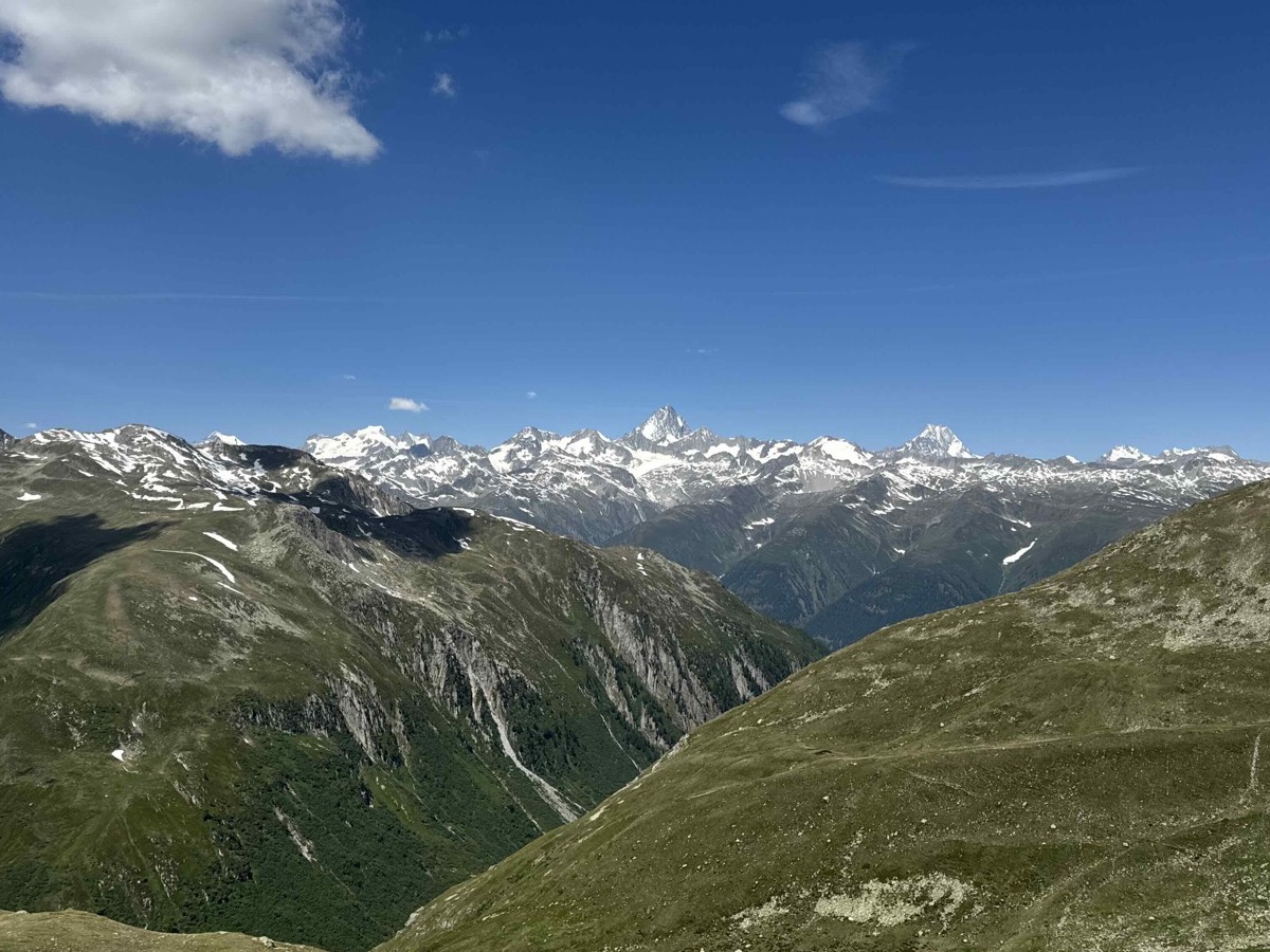 Panoramic view from the top of the Nufenenpass — green Alpine slopes dropping into a deep valley with the pass road visible in the lower right, a long horizon of snow-capped peaks across the middle distance, bright blue sky with scattered clouds and a patch of snow in the foreground
