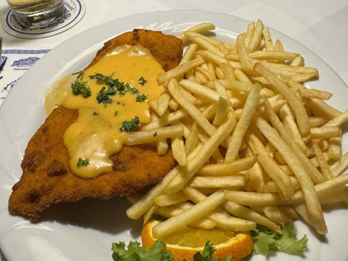A hearty plate of breaded schnitzel topped with a creamy sauce and parsley, a generous mountain of golden chips alongside, garnished with an orange slice and curly lettuce, a glass of Swiss beer in the background