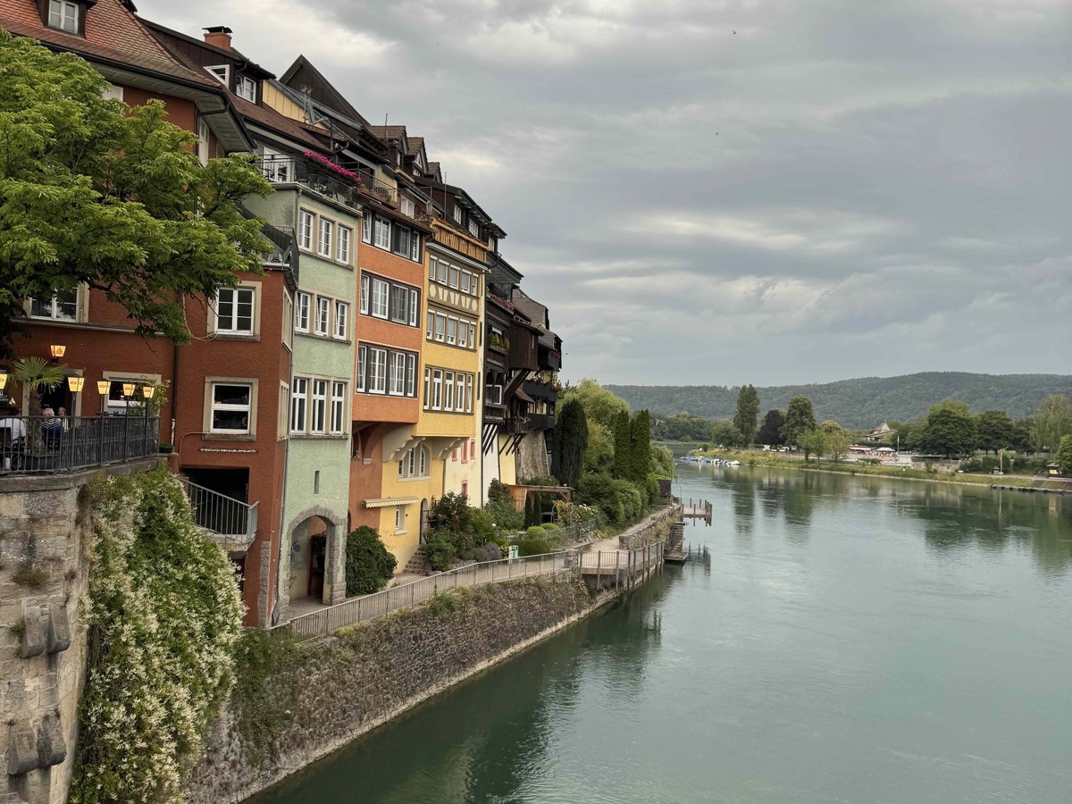 The old town of Laufenburg seen from across the Rhine — tightly packed cream, terracotta and yellow houses clustered along a steep riverbank above the curving green river, green hills rolling away in the distance under a moody grey sky