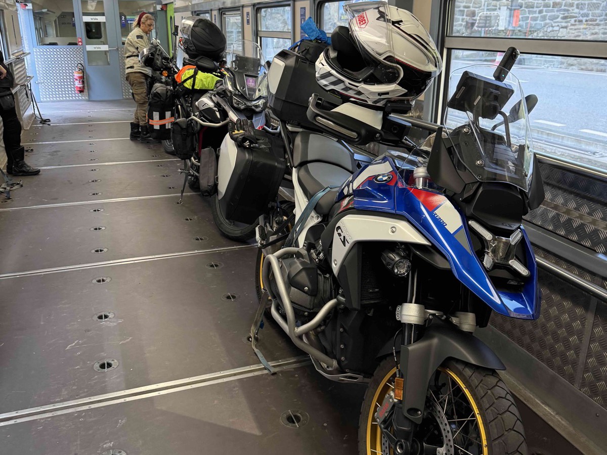 Motorbikes lined up inside the Lötschberg car-transporter train carriage — a blue, white and red BMW GS in the foreground with its helmet resting on the seat, more bikes lined up behind, a rider standing near the front of the carriage and metal anchor points in the floor