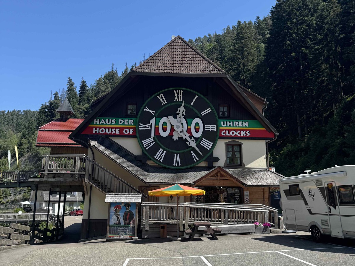 The 'Haus der 1000 Uhren / House of 1000 Clocks' in Triberg — a Black Forest chalet-style building with a giant green clock face mounted on the front and a tall pitched roof, a colourful umbrella over a picnic area in front, dark pine forest rising behind under a bright blue sky